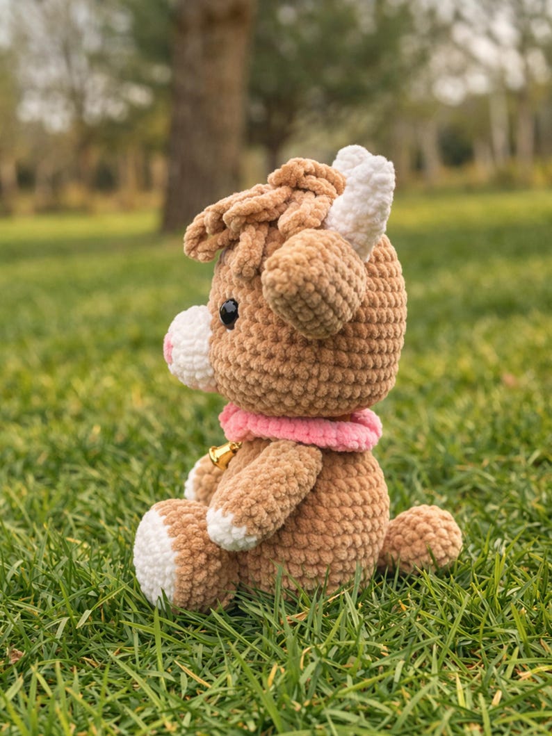 May include: A handmade crochet cow plush toy in shades of brown, white, and pink, sitting on green grass. The plush has a pink collar with a small gold bell. The cow's horns and hooves are white.