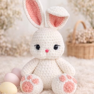 May include: A handmade white crochet bunny with pink inner ears, a pink nose, and black eyes. The bunny has pink paw pads and is sitting next to pastel-colored Easter eggs. The bunny is a soft, plush toy.