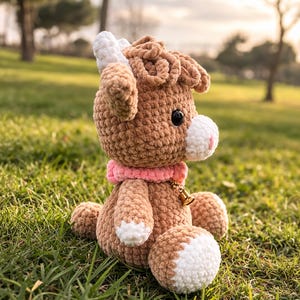 May include: A handmade crochet goat plushie in shades of brown and white, with a pink collar and a small gold bell. The plushie has black eyes and is sitting on green grass.