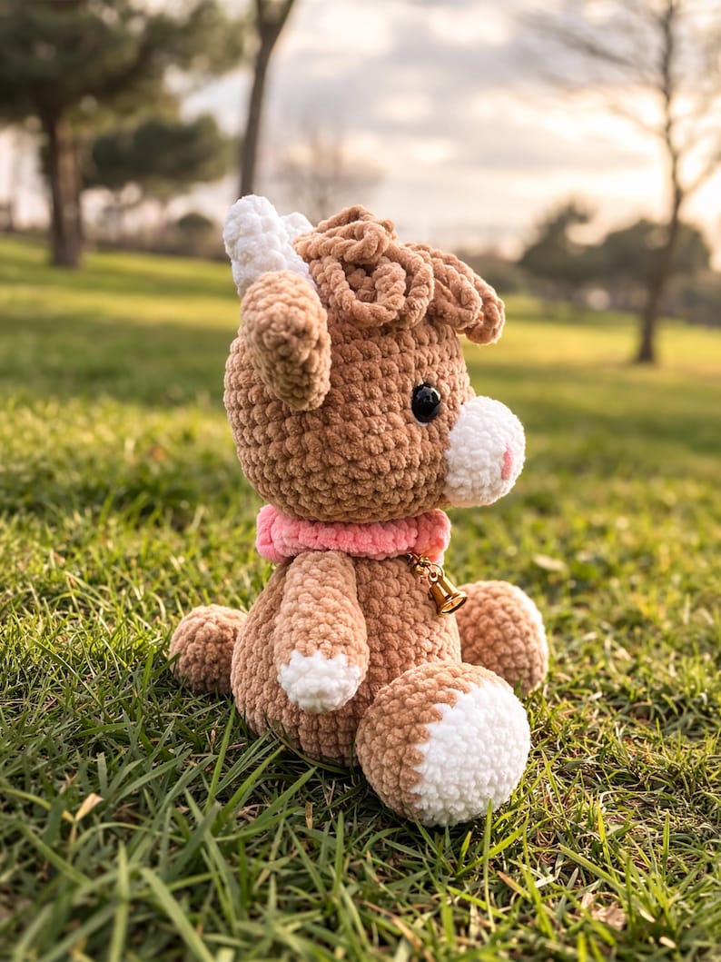 May include: A handmade crochet goat plushie in shades of brown and white, with a pink collar and a small gold bell. The plushie has black eyes and is sitting on green grass.