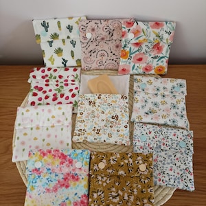 Peut inclure: Une collection de dix petites pochettes carrées en tissu avec fermetures à pression, présentant divers motifs floraux, botaniques et fruitiers. Une pochette est entrouverte, révélant un pain de savon rectangulaire. Les pochettes sont sur un tapis rond.