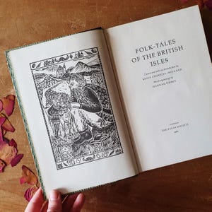 May include: An open book titled "FOLK-TALES OF THE BRITISH ISLES" with a black and white illustration on the left page. The illustration depicts a man and two figures in a landscape. The book's cover has a green and white patterned border.