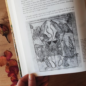 May include: An open book with a black and white illustration of a carriage being pulled by griffins. The illustration is from an old manuscript. The page also contains text in English.
