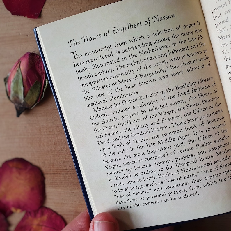 Puede incluir: Libro abierto con el t&iacute;tulo "The Hours of Engelbert of Nassau". El texto describe el origen del manuscrito en los Pa&iacute;ses Bajos a finales del siglo XV. El libro tiene una cubierta azul y est&aacute; sobre una superficie de madera.