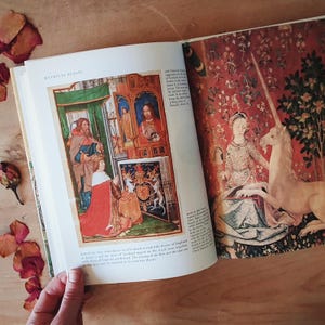 May include: An open book with illustrations of medieval art. One page features a colorful depiction of a royal ceremony, while the other showcases a tapestry with a woman and a unicorn. The book is titled "Mythical Beasts".