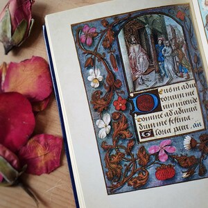 Puede incluir: Un libro abierto con una p&aacute;gina de manuscrito iluminado detallada. La p&aacute;gina presenta una colorida ilustraci&oacute;n de una escena hist&oacute;rica, rodeada de bordes florales y foliados. El texto de la imagen est&aacute; en lat&iacute;n. Capullos y p&eacute;talos de rosa secos sobre la superficie de madera.