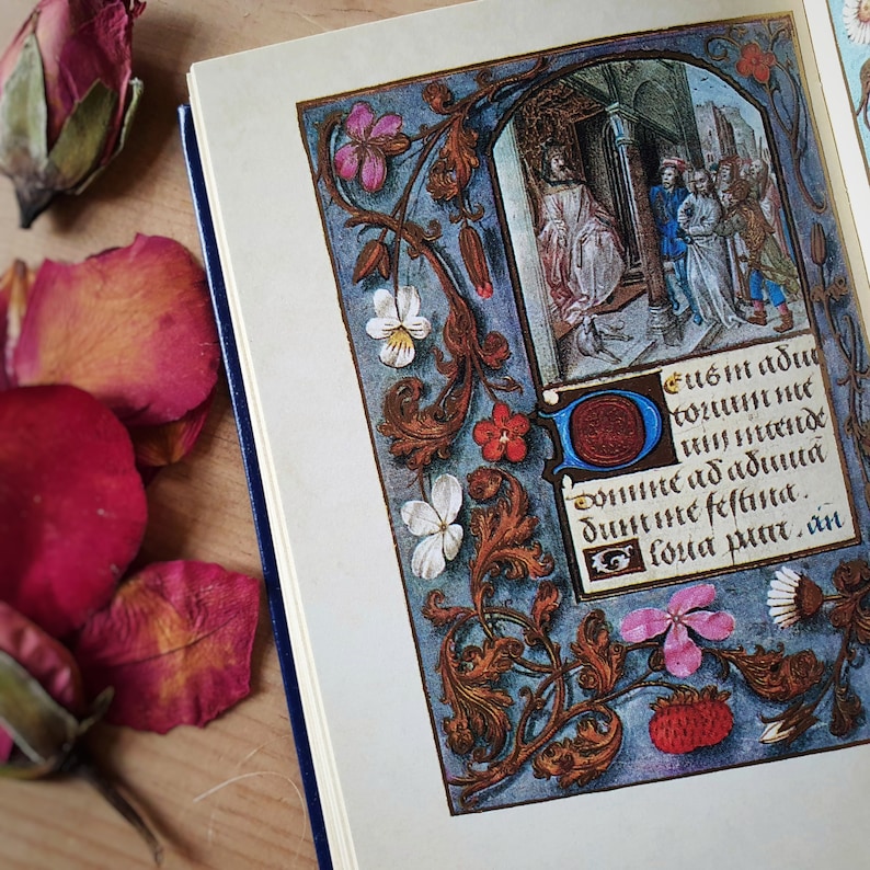 Puede incluir: Un libro abierto con una p&aacute;gina de manuscrito iluminado detallada. La p&aacute;gina presenta una colorida ilustraci&oacute;n de una escena hist&oacute;rica, rodeada de bordes florales y foliados. El texto de la imagen est&aacute; en lat&iacute;n. Capullos y p&eacute;talos de rosa secos sobre la superficie de madera.