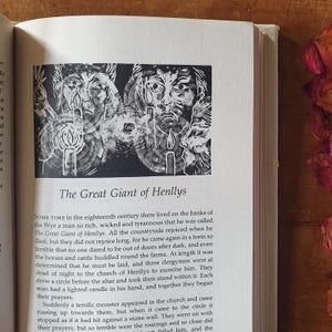 May include: An open book page with a black and white illustration of three faces with candles, and the title "The Great Giant of Henllys." The page is surrounded by text, and the book is on a wooden surface.