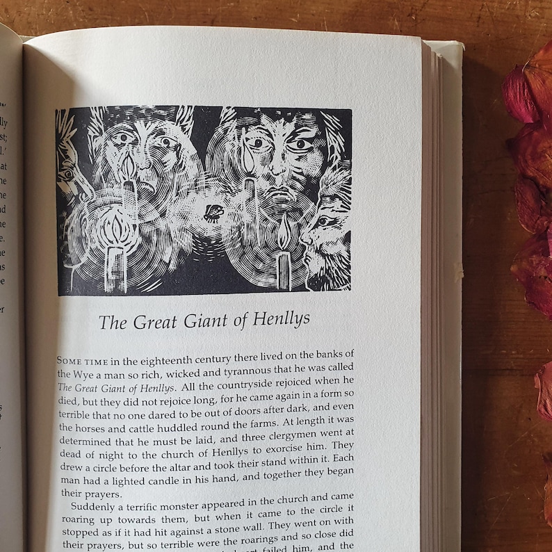 May include: An open book page with a black and white illustration of three faces with candles, and the title "The Great Giant of Henllys." The page is surrounded by text, and the book is on a wooden surface.