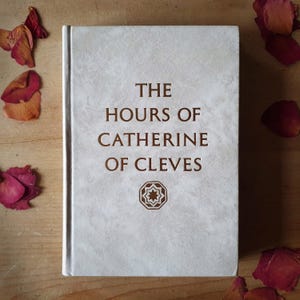 Może przedstawiać: Książka w twardej oprawie zatytułowana "THE HOURS OF CATHERINE OF CLEVES" w brązowych literach na jasnoszarej, teksturowanej okładce. Poniżej tytułu znajduje się geometryczny wz&oacute;r. Suche płatki r&oacute;ż są rozrzucone wok&oacute;ł książki na drewnianej powierzchni.