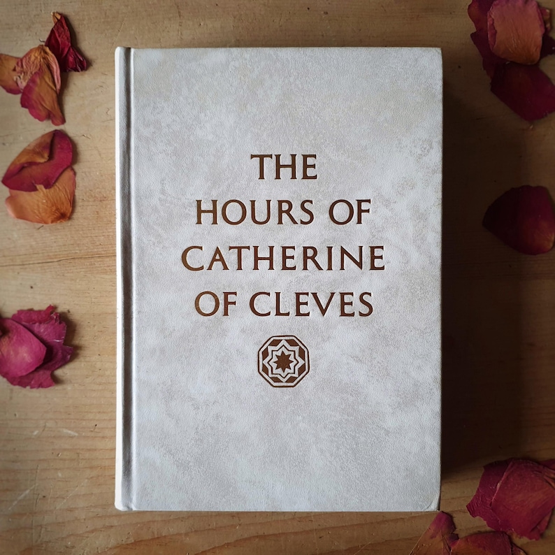 Może przedstawiać: Książka w twardej oprawie zatytułowana "THE HOURS OF CATHERINE OF CLEVES" w brązowych literach na jasnoszarej, teksturowanej okładce. Poniżej tytułu znajduje się geometryczny wz&oacute;r. Suche płatki r&oacute;ż są rozrzucone wok&oacute;ł książki na drewnianej powierzchni.
