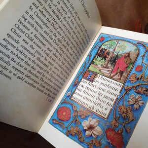 Puede incluir: Un libro abierto con una ilustraci&oacute;n detallada e iluminada. La obra de arte presenta una escena con figuras, una cruz y texto en un marco decorativo con motivos florales y foliares en azul, rojo y dorado. El texto est&aacute; en escritura g&oacute;tica.
