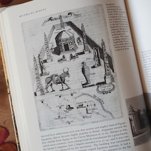May include: An open book displays a detailed black and white illustration of ancient Egyptian structures and figures. The image includes obelisks, a temple, a cow, and a map, with the title "MYTHICAL BEASTS" at the top.