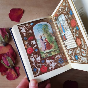 Puede incluir: Un libro abierto con ilustraciones y texto detallados. Las p&aacute;ginas presentan im&aacute;genes coloridas de figuras religiosas y bordes florales en rojo, azul y blanco. El libro tiene una cubierta azul oscuro y bordes dorados.