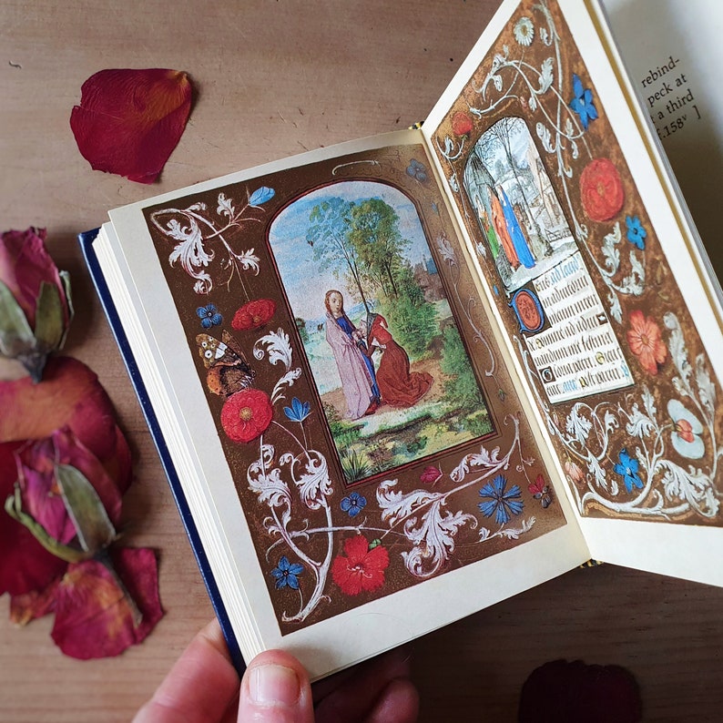 Puede incluir: Un libro abierto con ilustraciones y texto detallados. Las p&aacute;ginas presentan im&aacute;genes coloridas de figuras religiosas y bordes florales en rojo, azul y blanco. El libro tiene una cubierta azul oscuro y bordes dorados.
