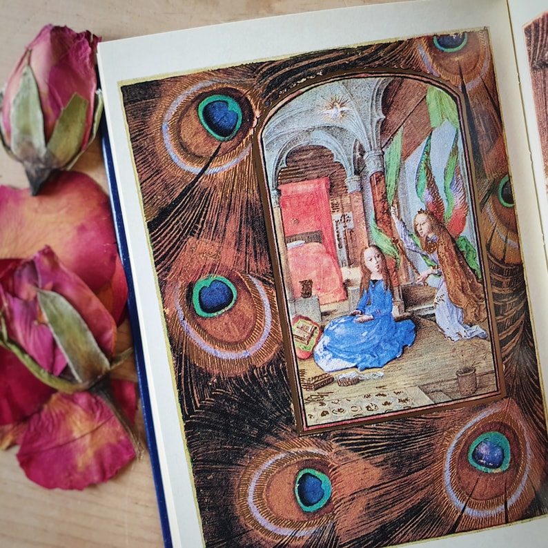 Puede incluir: Un libro abierto con una ilustraci&oacute;n medieval de una mujer y un &aacute;ngel, enmarcada por dise&ntilde;os de plumas de pavo real. P&eacute;talos y capullos de rosa secos est&aacute;n colocados cerca del libro. Borde dorado.
