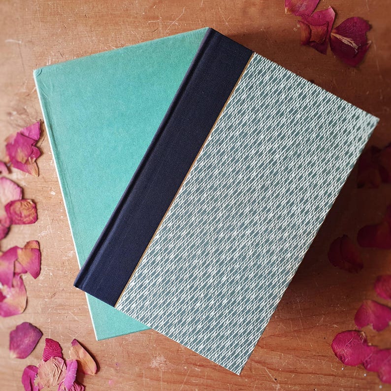 May include: Two hardcover books with teal and patterned covers. One book is open, revealing a teal cover and a navy blue spine. The other book has a light green cover with a white crisscross pattern. Dried rose petals are scattered around the books.