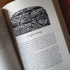 May include: An open book with the title "Croglin Grange" and a black and white illustration. The illustration depicts a building with a terrace and a distant view, framed by a geometric design. The text is in black font.