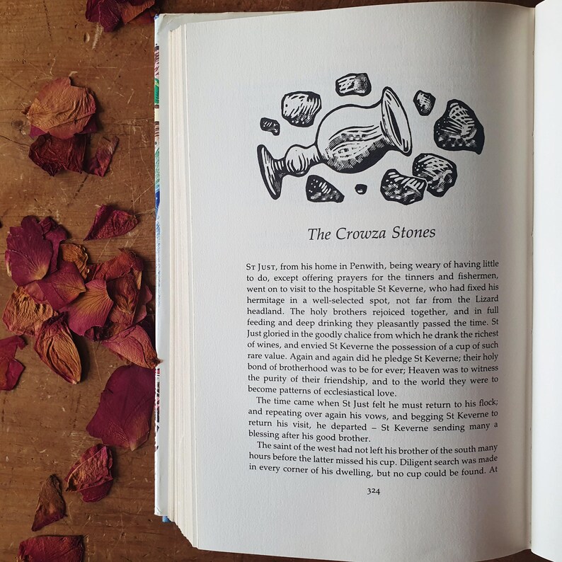 May include: Open book page with text and an illustration of a chalice and stones. The title "The Crowza Stones" is visible. Dried rose petals in shades of red and brown are scattered on a wooden surface.