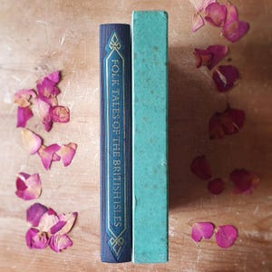May include: Two vintage books stand upright on a wooden surface, one navy blue with gold accents and the title "Folk Tales of the British Isles." The other book is a faded teal color. Scattered around are dried pink rose petals.