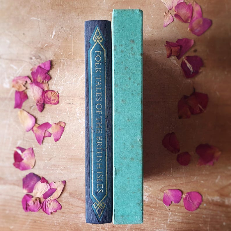 May include: Two vintage books stand upright on a wooden surface, one navy blue with gold accents and the title "Folk Tales of the British Isles." The other book is a faded teal color. Scattered around are dried pink rose petals.