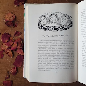 May include: Open book with text and an illustration of three women's heads in a decorative border. The title reads "The Three Heads of the Well." Dried rose petals are scattered on a wooden surface.