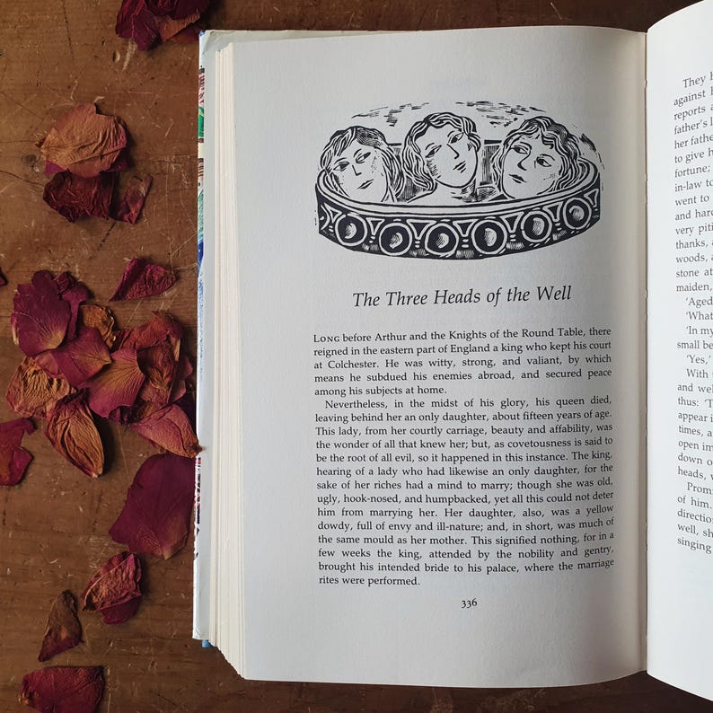 May include: Open book with text and an illustration of three women's heads in a decorative border. The title reads "The Three Heads of the Well." Dried rose petals are scattered on a wooden surface.