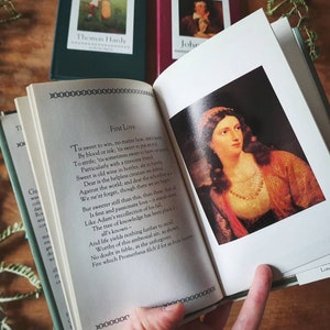 Puede incluir: Un libro abierto con un poema titulado "Primer amor" de Thomas Hardy. El poema est&aacute; impreso en una p&aacute;gina blanca con un borde negro. El libro est&aacute; abierto en una p&aacute;gina con una pintura de una mujer con cabello oscuro y un vestido rojo y dorado. La pintura est&aacute; enmarcada con un borde blanco. El libro est&aacute; sobre una mesa de madera con plantas verdes en el fondo.