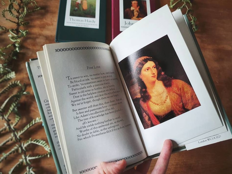 Puede incluir: Un libro abierto con un poema titulado "Primer amor" de Thomas Hardy. El poema est&aacute; impreso en una p&aacute;gina blanca con un borde negro. El libro est&aacute; abierto en una p&aacute;gina con una pintura de una mujer con cabello oscuro y un vestido rojo y dorado. La pintura est&aacute; enmarcada con un borde blanco. El libro est&aacute; sobre una mesa de madera con plantas verdes en el fondo.