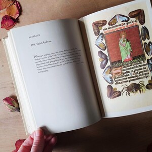 Może przedstawiać: Otwarta książka z tekstem "SUFFRAGE" i "119. Saint Ambrose." na lewej stronie. Prawa strona zawiera kolorową ilustrację postaci religijnej otoczonej ozdobnymi obramowaniami i tekstem. Suche płatki r&oacute;ż są rozrzucone wok&oacute;ł książki.