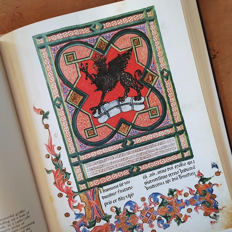 May include: An open book displaying a vibrant medieval illustration. The artwork features a black griffin atop a white bale, set against a red background within an ornate frame. The page is adorned with colorful floral and geometric patterns, and handwritten text.