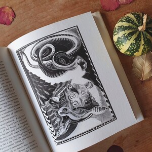 May include: An open book with a detailed black and white illustration of a snake and a turtle. The illustration is framed with a decorative border. A small decorative pumpkin and leaves are on the wooden surface.