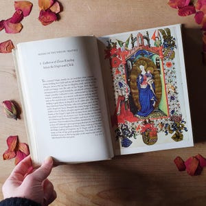 Może przedstawiać: Otwarta książka z szczeg&oacute;łową ilustracją Matki Boskiej z Dzieciątkiem, otoczona ozdobnymi obramowaniami. Na przeciwległej stronie znajduje się tekst zatytułowany "HOURS OF THE VIRGIN-MATINS". Suche płatki r&oacute;ż są rozrzucone wok&oacute;ł książki.