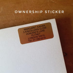 May include: A rectangular, bronze-colored ownership sticker with rounded corners is affixed to a white surface. The sticker features the text "OWNERSHIP STICKER" at the top, and below it, the name "MARGARET KAHAN" and an address and phone number.