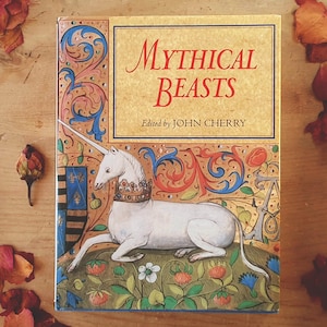 May include: A hardcover book titled "Mythical Beasts" edited by John Cherry. The cover features a white unicorn with a golden crown, set against a backdrop of floral and ornate designs in red, blue, and gold. The book is on a wooden surface.