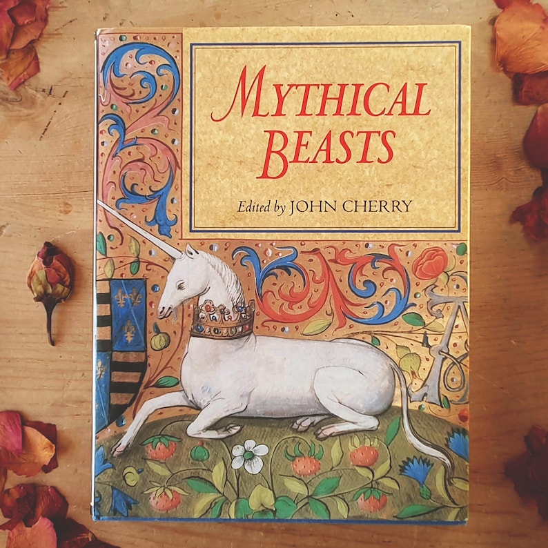 May include: A hardcover book titled "Mythical Beasts" edited by John Cherry. The cover features a white unicorn with a golden crown, set against a backdrop of floral and ornate designs in red, blue, and gold. The book is on a wooden surface.