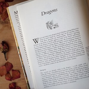 May include: Open book with the title "Dragons" and a detailed illustration of a dragon. The pages are cream-colored, and the text is in black. Dried rose petals are scattered around the book on a wooden surface.