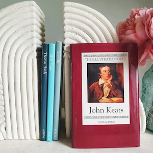 Puede incluir: Un conjunto de cuatro libros con lomos rojos, azules y verdes. Los libros se titulan "Lord Byron", "W.B. Yeats", "Thomas Hardy" y "John Keats: The Illustrated Poets". Los libros est&aacute;n en un estante blanco con sujetalibros blancos con forma de arco&iacute;ris.