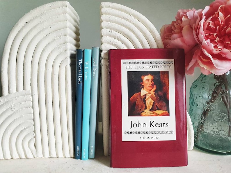Puede incluir: Un conjunto de cuatro libros con lomos rojos, azules y verdes. Los libros se titulan "Lord Byron", "W.B. Yeats", "Thomas Hardy" y "John Keats: The Illustrated Poets". Los libros est&aacute;n en un estante blanco con sujetalibros blancos con forma de arco&iacute;ris.