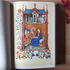 Może przedstawiać: Szczeg&oacute;łowa ilustracja z manuskryptu. Scena religijna przedstawia kobietę w niebieskiej szacie z aureolą, czytającą. Anioł w pomarańczowym i niebieskim prezentuje zw&oacute;j. Scena jest obramowana elementami architektonicznymi i kwiatowymi.