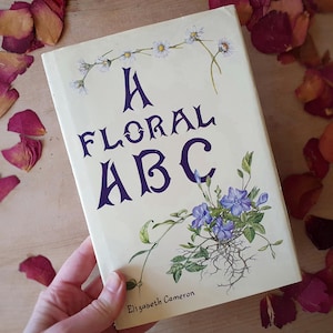 May include: A book titled "A Floral ABC" with a cream-colored cover. The title is in dark purple, with floral illustrations of daisies and blue flowers. The author's name, Elizabeth Cameron, is at the bottom.