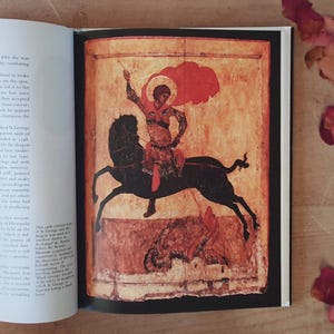 May include: An illustration from a book depicts a figure on a black horse, holding a spear. The rider is clad in red and gold, with a red halo. The background is a faded orange, with text visible on the left page.
