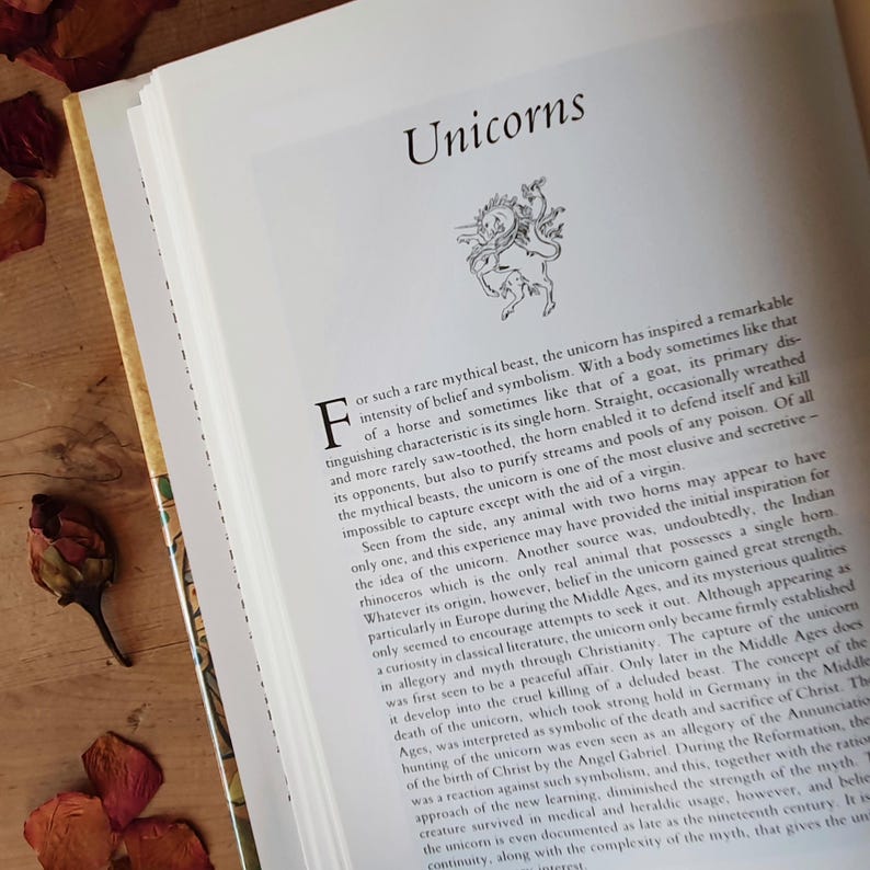 May include: Open book with the title "Unicorns" and an illustration of a unicorn. The pages are white with black text. Dried flower petals and a dried flower bud are on a wooden surface.