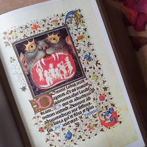 Może przedstawiać: Otwarta strona książki z ilustracją średniowieczną. Grafika przedstawia pysk potwora z postaciami w środku, otoczony ozdobnymi kwiatowymi obramowaniami. Widoczny jest r&oacute;wnież tekst łaciński.