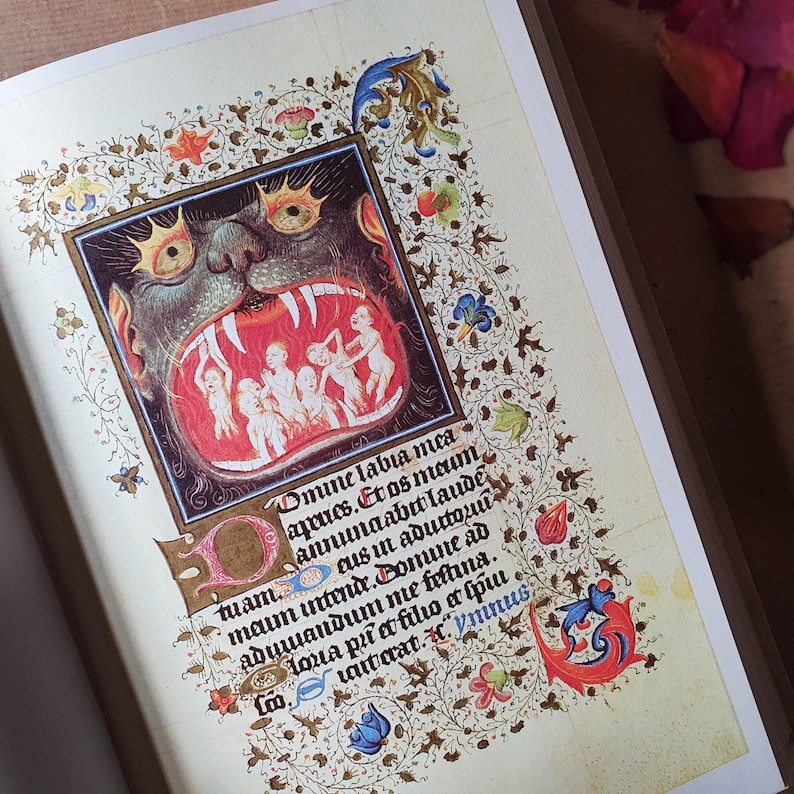Może przedstawiać: Otwarta strona książki z ilustracją średniowieczną. Grafika przedstawia pysk potwora z postaciami w środku, otoczony ozdobnymi kwiatowymi obramowaniami. Widoczny jest r&oacute;wnież tekst łaciński.