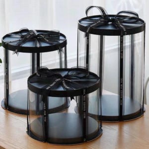 May include: Three clear, cylindrical cake boxes with black bases and lids. Each box is tied with a black ribbon printed with "BEST WISHES FOR YOU". The boxes are different heights, creating a tiered display.