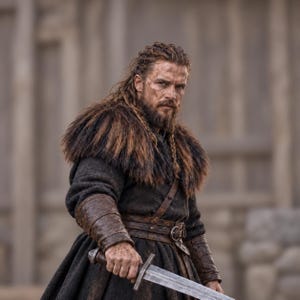 May include: A person with long, braided brown hair and a beard, wearing a dark, textured wool tunic with a thick, dark brown fur collar. The attire includes brown leather bracers and a leather belt with a metal buckle, while holding a steel sword.