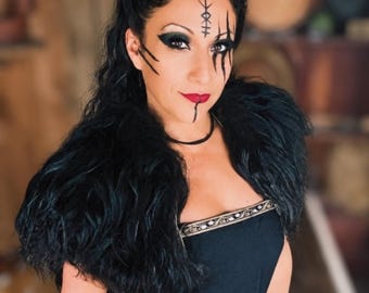 May include: A black sleeveless dress featuring a metallic silver embroidered trim along the neckline and waist, paired with a voluminous black faux fur shoulder wrap. The look includes dark eye makeup and black facial markings on the forehead and cheeks.