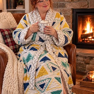 May include: A quilted robe featuring a geometric pattern of triangles in shades of mustard yellow, teal, and navy blue. The robe has a shawl collar and a matching tie belt, both decorated with a grey and white chevron print on a white background.