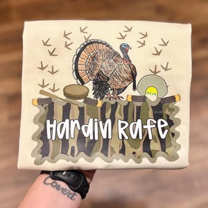 Può includere: T-shirt beige con un'illustrazione di tacchino, impronte e la scritta "Hardin Rafe" in bianco. Il tacchino è marrone, nero e arancione. Il testo è su uno sfondo mimetico.